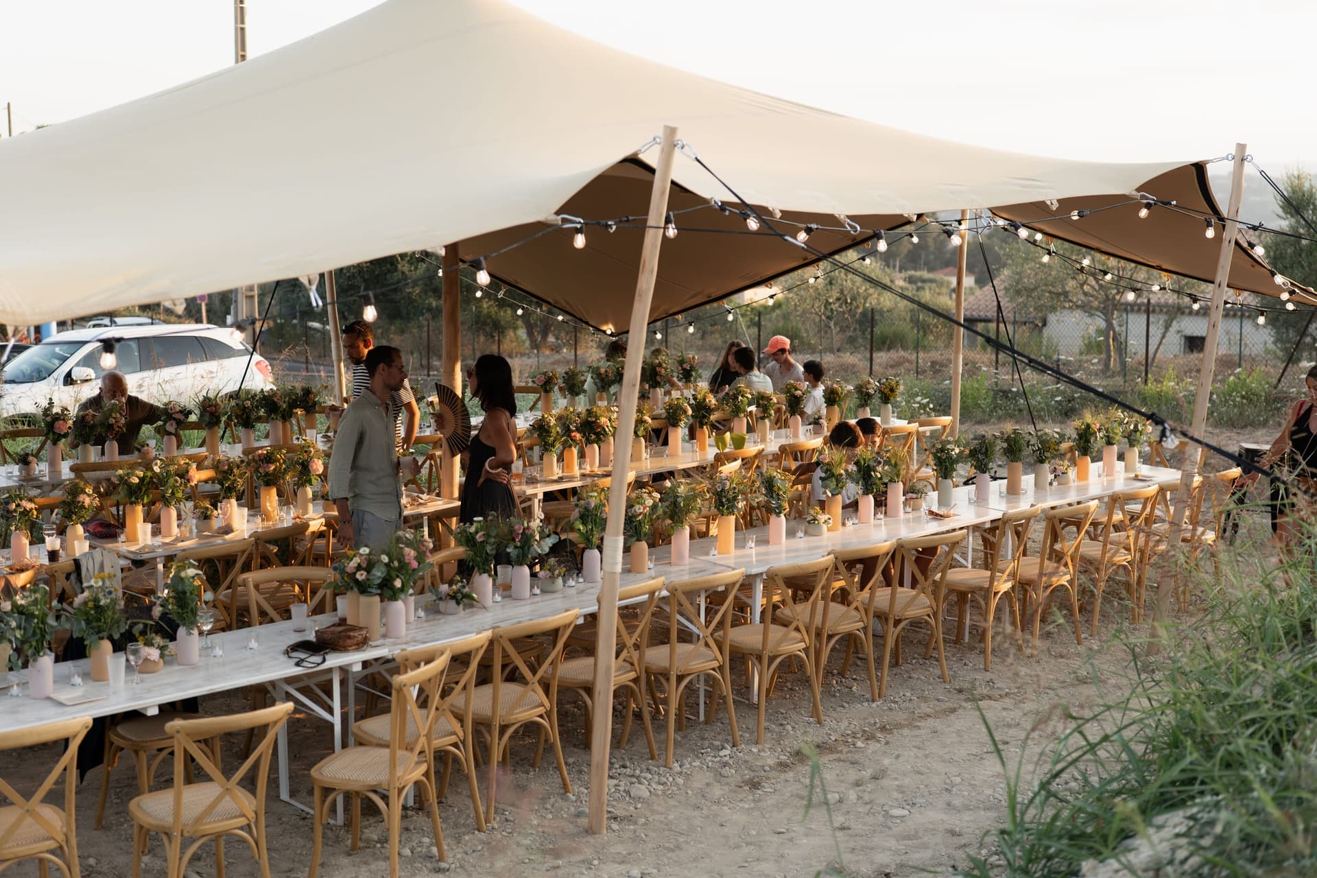 Longues tables de banquet dressées sous une tente stretch, à la lumière du soir, guirlandes guinguette allumées