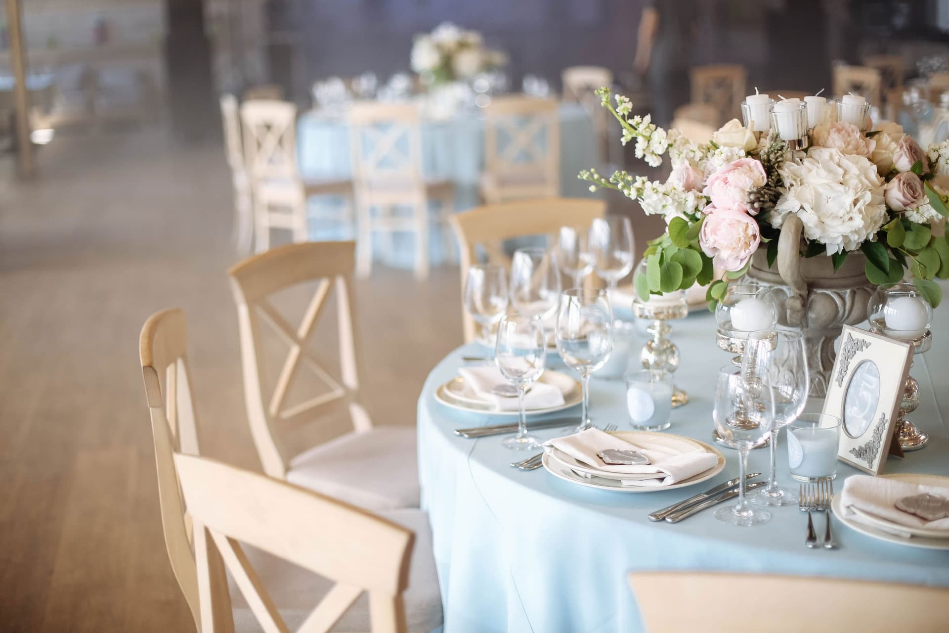 Table ronde dressée : verres à pied, couverts, assiettes et bouquet de pivoines sur une nappe bleu clair