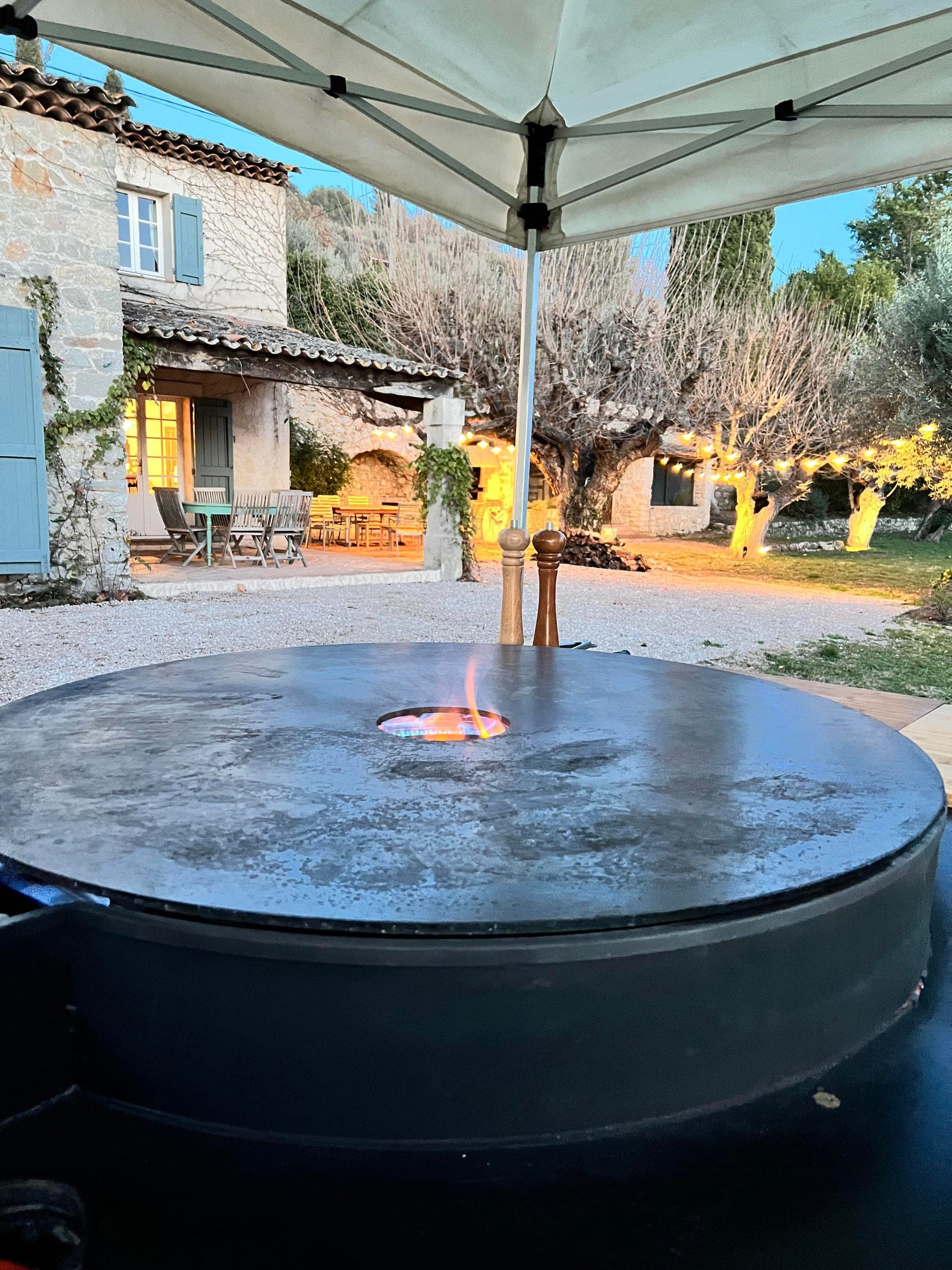 Plancha ronde allumée sous une tonnelle, devant une bastide provençale éclairée au crépuscule
