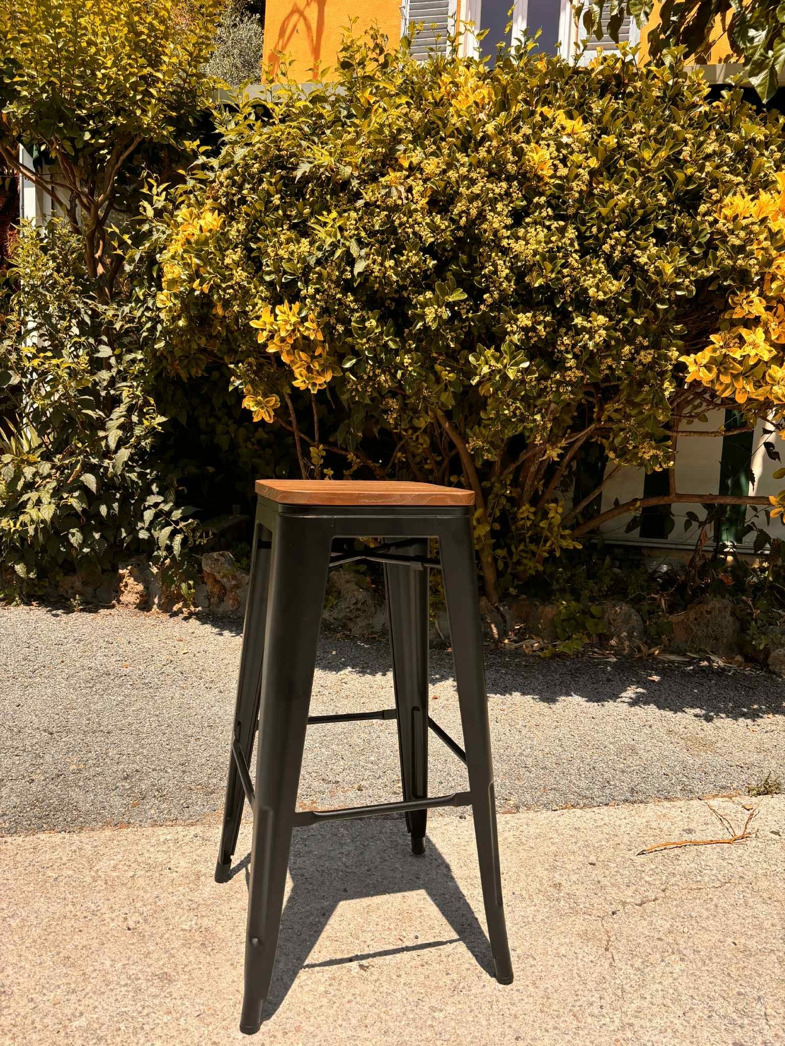Tabouret de bar en métal noir à assise de bois, posé au soleil devant un massif fleuri
