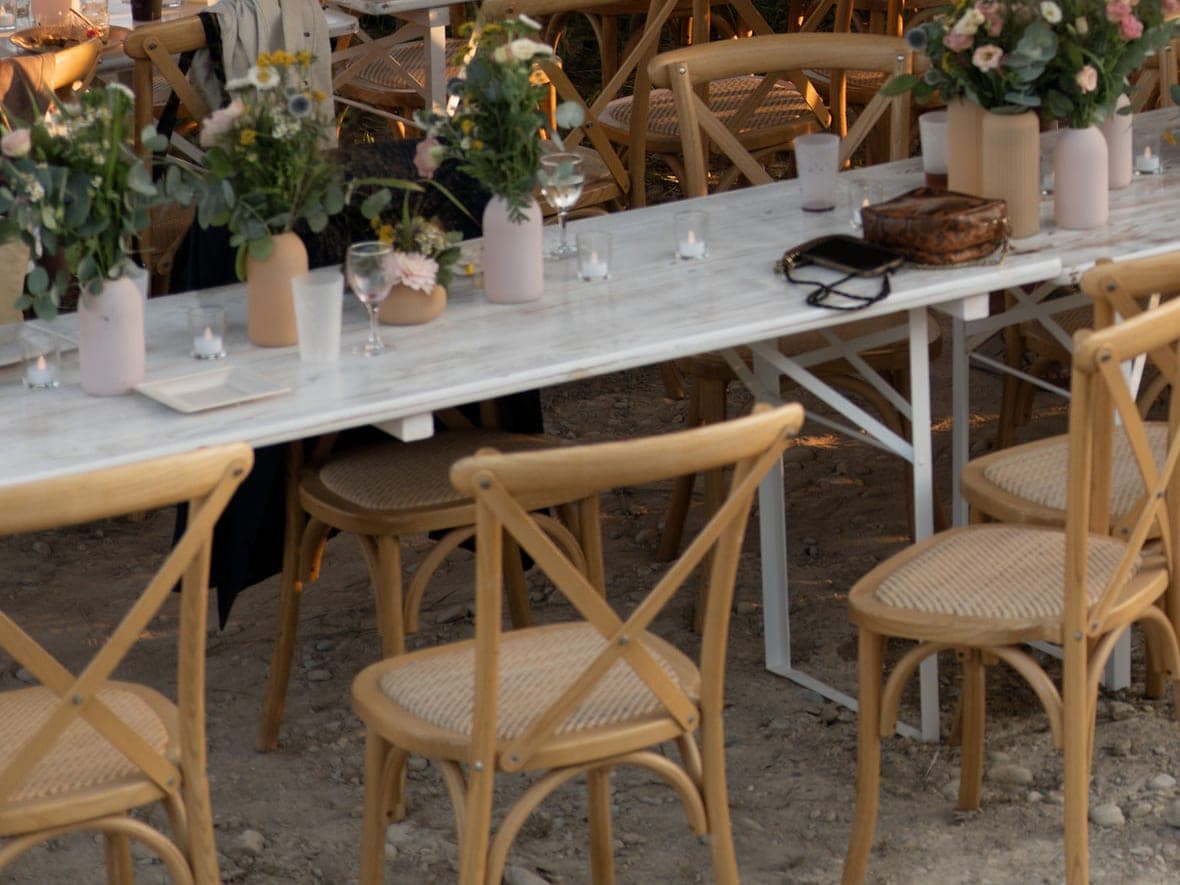 Tables de banquet dressées — verres, bougies et bouquets — et rangées de chaises bistrot en bois clair, à la lumière du soir